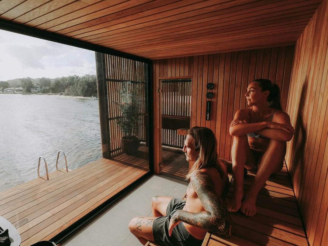 Two people relaxing in a timber sauna that opens onto the water